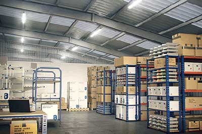 Binnenkant van een grote opslagunit met stellingen, planken vol dozen, een werktafel met laptop en pallets met dozen.