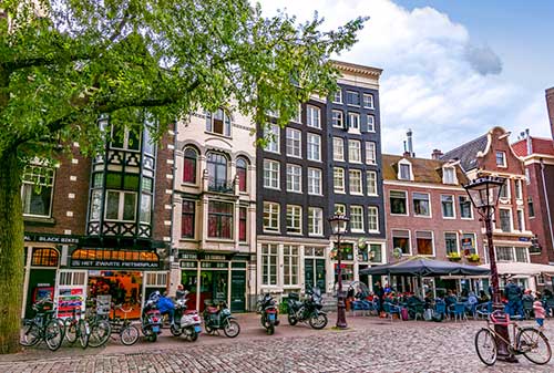 Urban street in the Netherlands with tall row houses, bicycles, cobblestone road, and ground-floor cafés and shops.